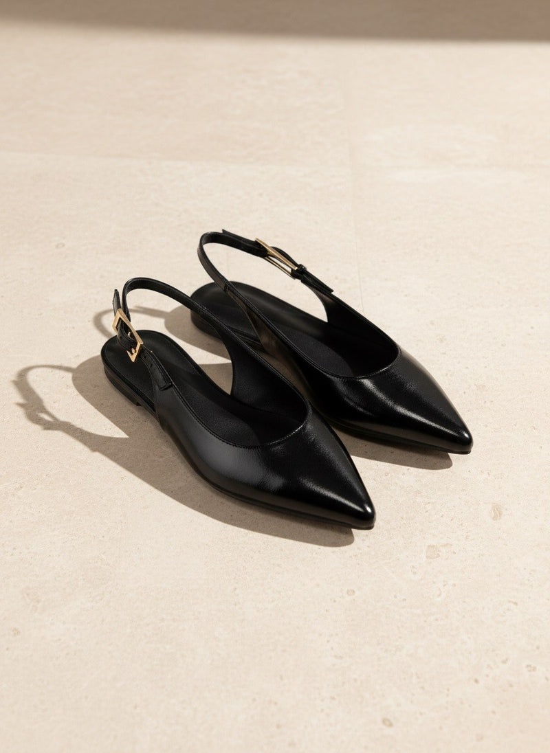 A pair of black leather pointed-toe slingback flats with gold-tone buckle details, displayed on a light beige stone surface.